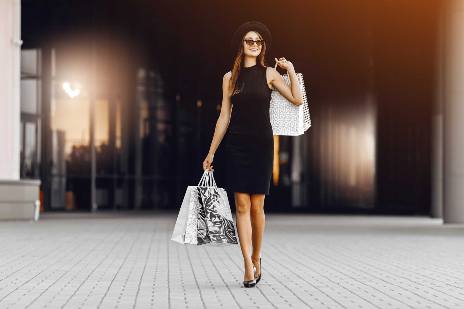 confident, happy, attractive young woman in a black dress and hat, wearing dark glasses, holding shopping bags in front of a shopping center. Black Friday, shopping